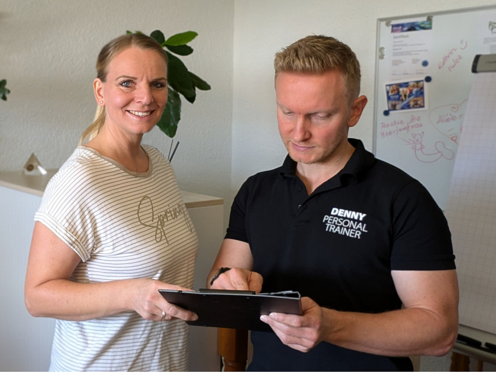 Personal Trainer Denny zeigt einer Kundin etwas auf einem Klemmbrett.