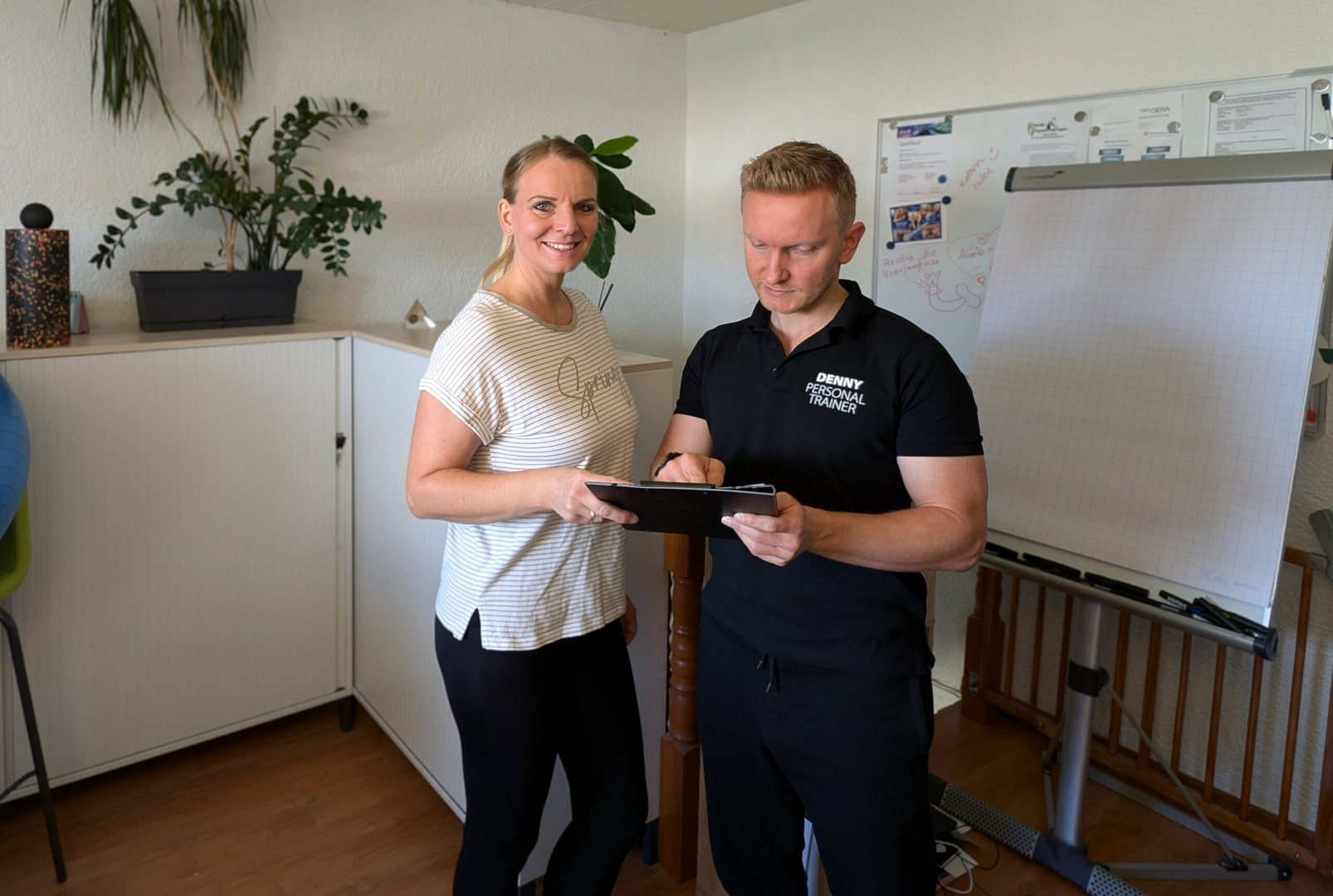 Personal Trainer Denny zeigt etwas auf einem Klemmbrett einer Frau. Beide stehen in einem Büro. Zudem stehen eine Faszienrolle und ein Faszienball auf dem Schrank.