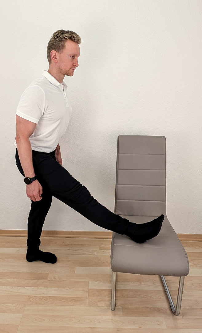 Denny steht aufrecht, ein Stuhl steht vor ihm. Das rechte Bein legt er gestreckt auf dem Stuhl vor ihm ab. Sein Oberkörper ist leicht vorgeneigt.