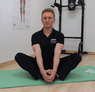 Denny sitzt auf dem Boden. Die Fußsohlen berühren einander. Die Knie gehen zur Seite nach unten.