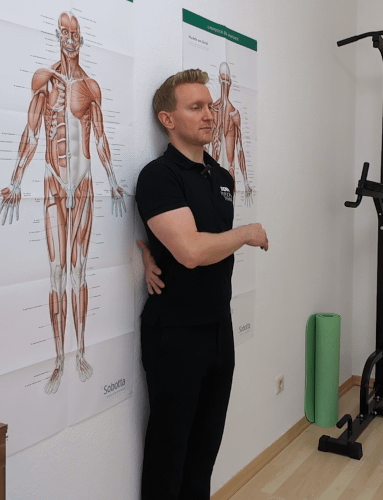 Hohlkreuz Selbsttest - Wandtest: Denny steht mit dem Rücken an der Wand. Er führt seinen linken Arm zwischen Wand und unteren Rücken durch.