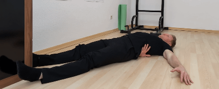 Boden-Liegetest: Denny liegt auf dem Rücken am Boden. Er führt seinen rechten Arm zwischen unterem Rücken und Boden durch.