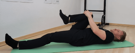 Denny liegt mit dem Rücken am Boden. Ein Bein ist gestreckt, das andere Bein ist mit dem Knie zur Brust gezogen.