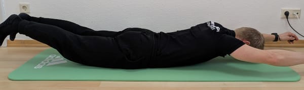 Denny ist in Bauchlage. Arme und Beine sind gestreckt und vom Boden angehoben.
